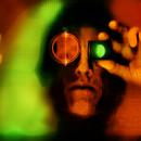 A color self-portrait scanograph representing disintegration of  the body and vision.  The primary colors are green, red and black. My face is looking straight ahead and I am holding a monocular in front of my eye. The sides of my face are silhouetted in black. The image is very distorted and phased. This image is from my “Transmog Project” body of work. 