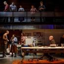 On the stage, there are six people starting with a whilte male in red shirt, a white male with brown cardigan looking at a black female as they talked along with a white female with curly hair looking at them,  An older male sitting by a table with a laptop in front of him looking at them and an older woman standing in the corner. 