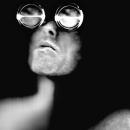A Black & White self-portrait scanograph from my “Blind Vision” series. In this image I am shown head and shoulders with face looking up on a black background. Over my eyes are large magnification loupes.  The loupes are the size of teacups and give me an almost robotic look. 