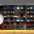 A large sculpture made of many rectangular cubbies filled with shaped acoustic foam stands flat against a wall. The depth of the cubbies casts shadows over flat red, blue, yellow, aqua, and pink foam panels. Flat, black pieces of wood in various shapes cover parts of the sculpture. The left side has a half circle cut out with a musical triangle hanging inside.