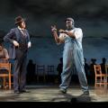 A black man in blue overalls is signing and a while male in a suit looking at the black male in overalls.