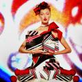 A photomontage of a female model sitting cross-legged on the floor holding a gift-wrapped box. The holiday gift wrap is mimicked in the models clothing. The background is filled with colorful festive designs. This image is from my “Neo Glam” series of reimagined fashion portraits.