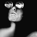 A Black & White self-portrait scanograph from my “Blind Vision” series. In this image I am shown head and shoulders with face looking up on a black background. Over my eyes are large magnification loupes.  The loupes are the size of teacups and give me an almost robotic look. 