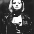 A Black & White photographic portrait of a female model.  She is wearing a black hair band pushing back her platinum blond hair. She is wearing a black leather jacket which is open revealing some of her torso. She is holding a mask near her chin. It is attached to a stick. The studio light from above is casting a shadow of the mask onto her torso. The shadow looks like a butterfly.