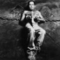 A photographic portrait of a young Latino. He is laying on a fabric background. He is shirtless and holding a crucifix over his torso. Behind him are angel’s wings which are the same color as the background fabric. His eyes are closed and a beautiful white light is illuminating his wings and body. This image is from my “Remember – an AIDS memorial retrospective.” Body of work.