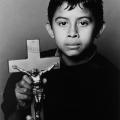 A Black & White photographic portrait of a young Latin boy holding a crucifix. He is staring directly into the camera with beautiful dark eyes. He has a somber expression. Both of his parents have died from AIDS.  This portrait is from my “Remember – an AIDS memorial Retrospective” body of work. 