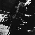 A Black & White photographic portrait of a young man seated on a chair in front of a black wall. He is wearing a black sport coat and holding piano pedals in his hands. He is looking off camera to the right side of the frame. He has a small pin in the shape of a guitar affixed to his lapel The image is photographed on the diagonal creating tension within the frame. On the white concrete floor to his left is a vintage metal circular fan. This image is from my “Remember – an AIDS Memorial Retrospective” body 