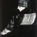 A Black & White photographic portrait of a young woman seated on a chair. The woman and chair is turned with their backs toward the camera.  The woman has her face turned so you can see her profile. She is wearing a silvery fabric gown. The same fabric is wrapped around her head as a hair piece. She has black batiste fabric draped over her shoulders and running down her left arm. The style of the gown and pose resembles an image that would have appeared in a 1920’s Vogue magazine.